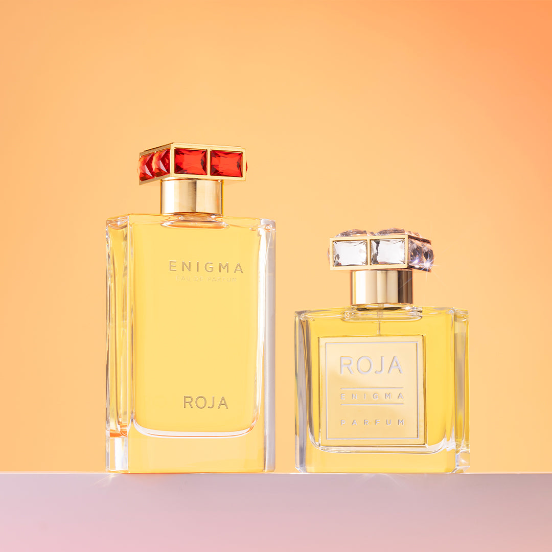 Two clear glass perfume bottles filled with golden liquid, labeled ENIGMA ROJA, stand side by side on a light surface against a warm orange gradient background. One bottle is taller with a red-accented cap, the other shorter with a clear cap.