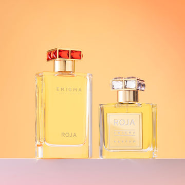Two clear glass perfume bottles filled with golden liquid, labeled ENIGMA ROJA, stand side by side on a light surface against a warm orange gradient background. One bottle is taller with a red-accented cap, the other shorter with a clear cap.