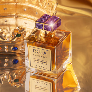 A bottle of Roja Parfums Great Britain perfume with a gold label and a gem-encrusted purple cap sits in front of an ornate, jeweled gold crown on a reflective gold surface.