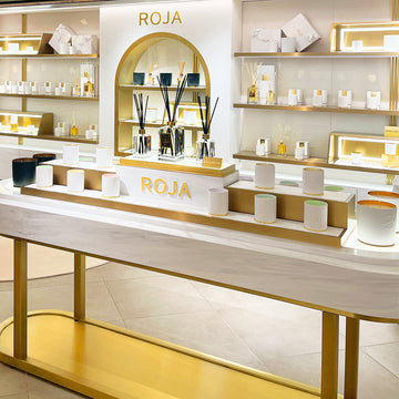 A luxurious fragrance store display featuring candles, diffusers, and boxed products on white and gold shelves with the brand name “ROJA” prominently shown. The setup is elegant and well-lit.