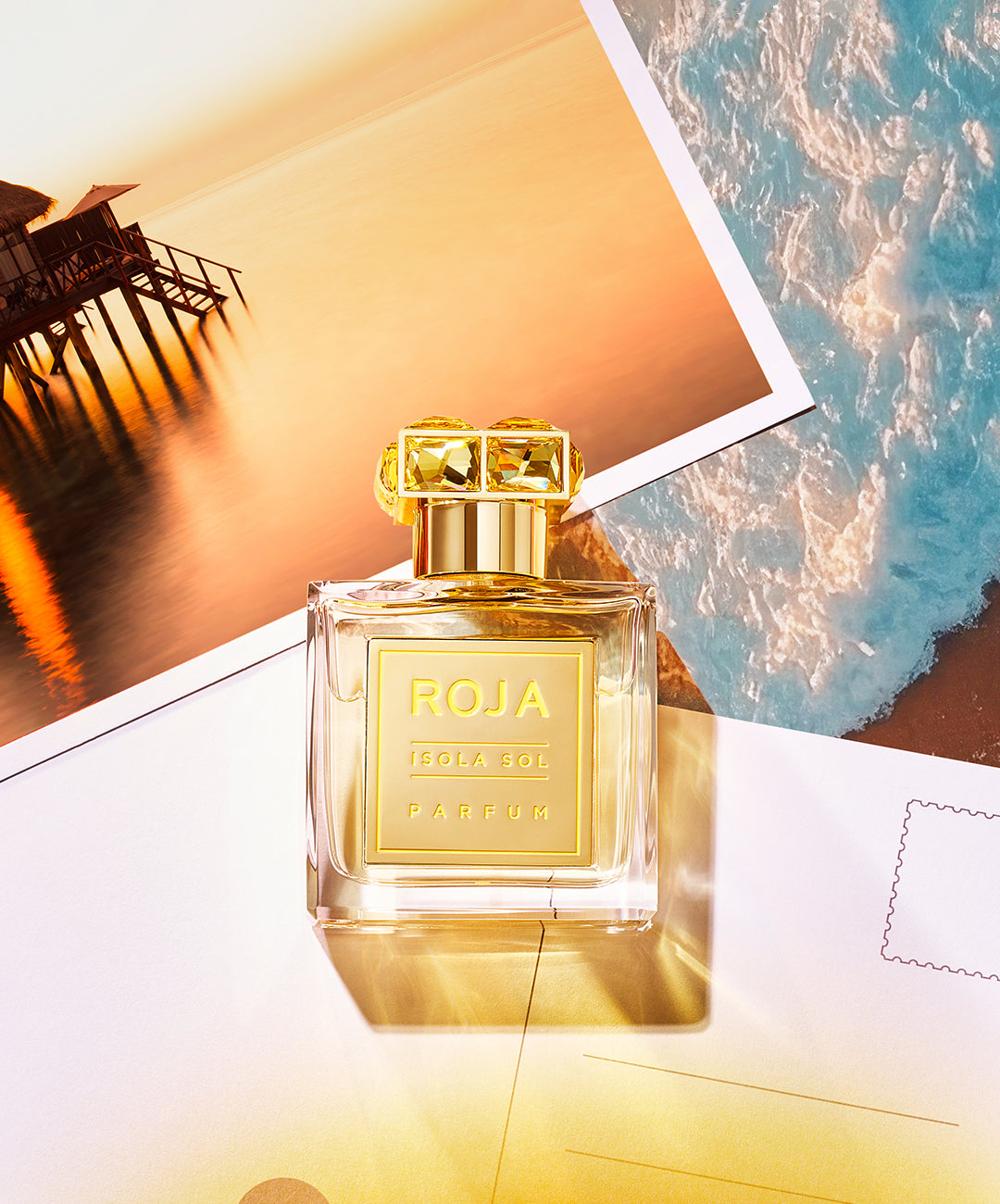 A bottle of Roja Isola Sol perfume sits on top of two postcards, one showing a tropical sunset over water, the other an aerial view of a beach with turquoise waves. The perfume bottle is golden and elegant, glowing in the sunlight.