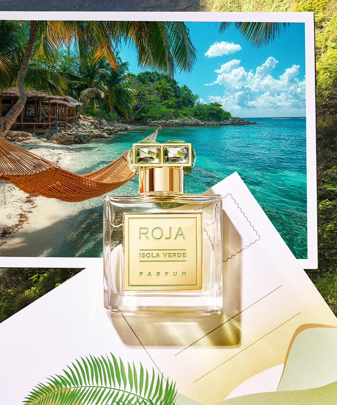 A bottle of Roja Isola Verde Parfum sits on a paper with palm leaf designs, in front of a scenic tropical beach photo with turquoise water, palm trees, and an orange hammock.