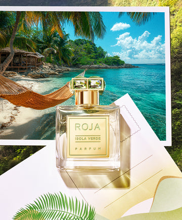 A bottle of Roja Isola Verde Parfum sits on a paper with palm leaf designs, in front of a scenic tropical beach photo with turquoise water, palm trees, and an orange hammock.