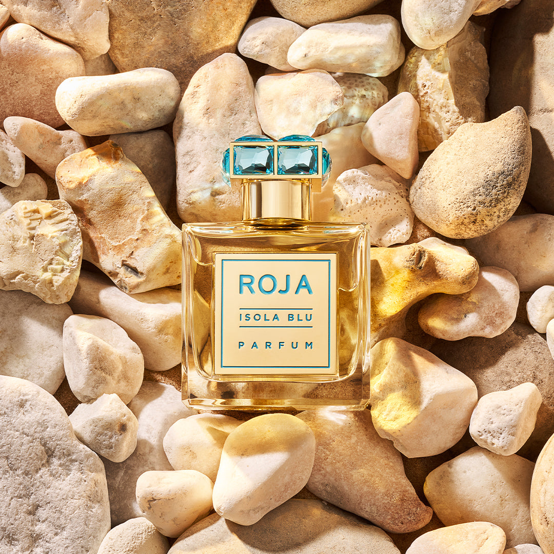 A bottle of Roja Isola Blu Parfum with a blue gem-like cap is placed on a bed of smooth, light-colored rocks. The label on the bottle is blue and gold.