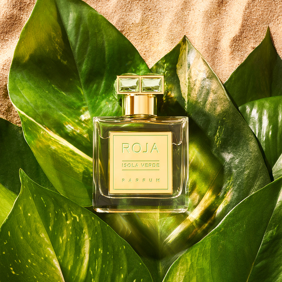 A bottle of ROJA Isola Verde Parfum rests on large green tropical leaves, set against a sandy background, evoking a fresh, luxurious, and exotic atmosphere.