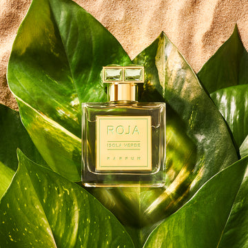 A bottle of ROJA Isola Verde Parfum rests on large green tropical leaves, set against a sandy background, evoking a fresh, luxurious, and exotic atmosphere.