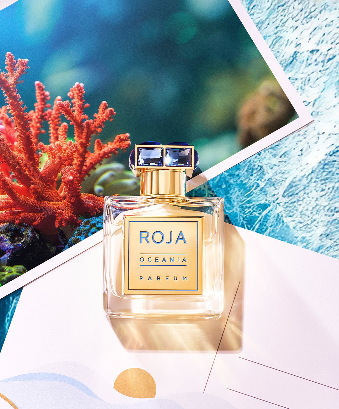 A bottle of Roja Oceania Parfum sits on top of postcards, one featuring vibrant coral and an underwater scene. The clear bottle has a gold label and cap, surrounded by soft, natural light.