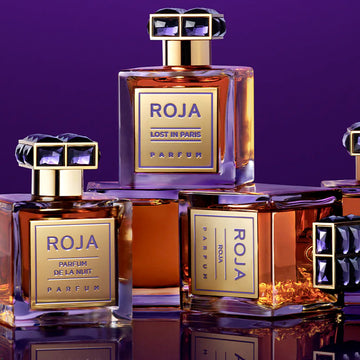 Four luxurious Roja Parfums bottles with gold labels and ornate black and gold caps are displayed against a purple background. The fragrances include Lost in Paris and De La Nuit.