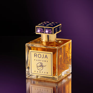A square glass bottle of golden Roja Parfums perfume with a gold and navy cap, set against a dark purple gradient background. The label on the bottle features gold and black detailing and a signature design.