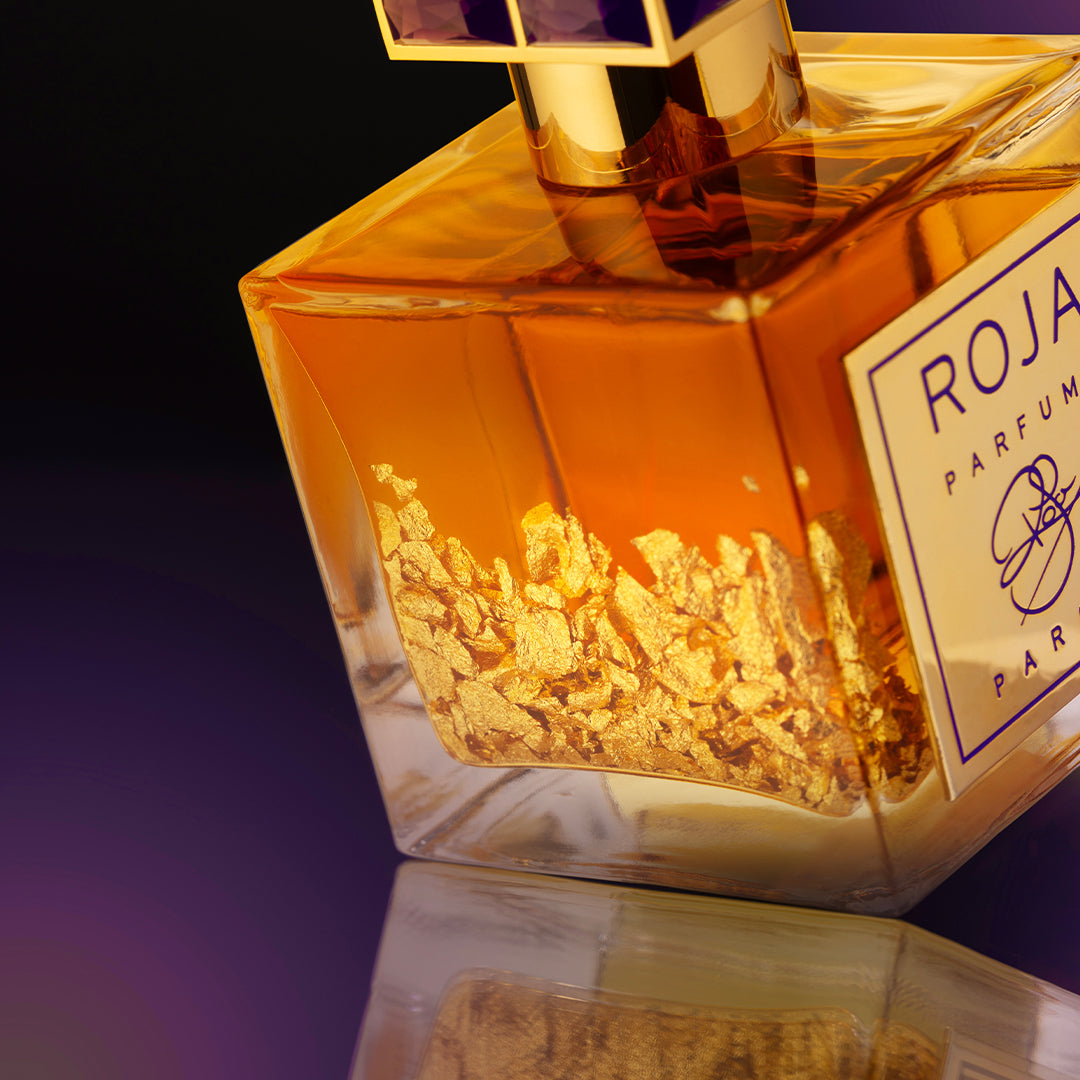 A close-up of a luxurious perfume bottle filled with amber liquid and visible gold flakes, labeled ROJA PARFUMS, set against a gradient purple and black background.