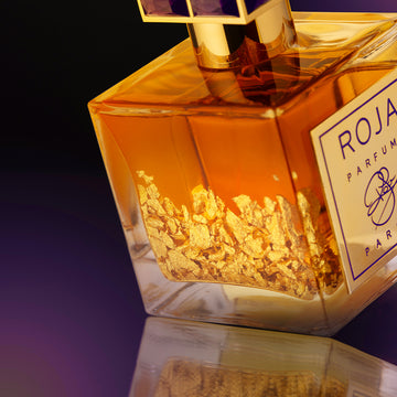 A close-up of a luxurious perfume bottle filled with amber liquid and visible gold flakes, labeled ROJA PARFUMS, set against a gradient purple and black background.