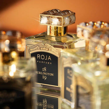 A close-up of a luxurious Roja Parfums bottle labeled 18 Burlington 19, featuring a gold and black design with a crystal-like cap, surrounded by other elegant perfume bottles.