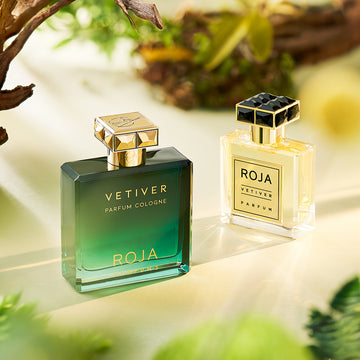 Two elegant perfume bottles labeled ROJA VETIVER placed on a light surface with a green, nature-inspired background. One bottle is green and gold, the other clear and gold, surrounded by leaves and branches.