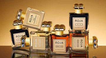 A group of luxurious Roja Parfums bottles with gold caps are artfully arranged against a gold gradient background, with reflections visible on the shiny surface below.