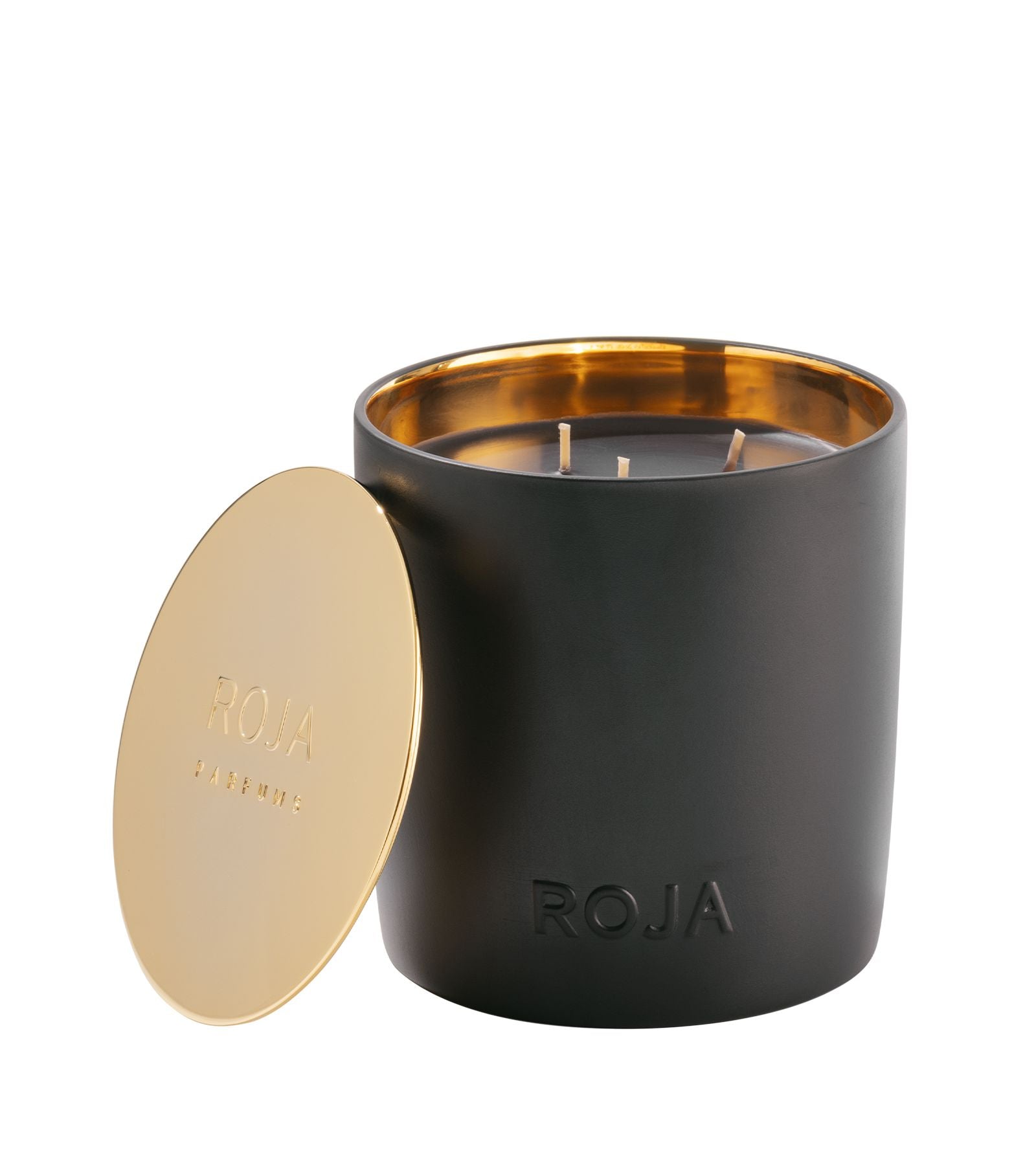A luxurious black candle holder from Roja Parfums, featuring an Amber Aoud candle, radiates olfactory majesty. Its gold interior with dual wicks and the embossed Roja name enhance its elegance. Nearby, the golden lid is engraved with sophistication.