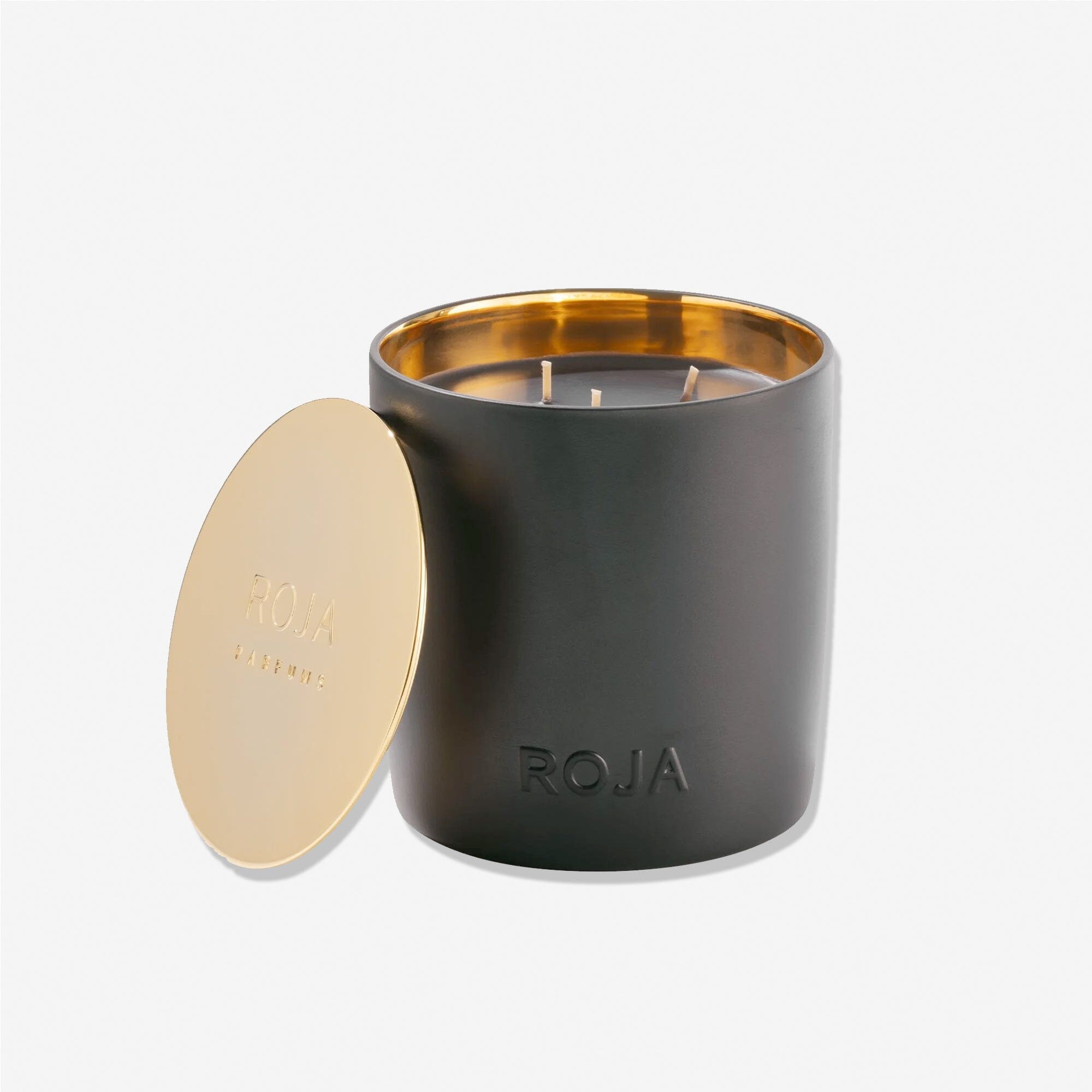 A matte black candle jar with Roja Parfums embossed on the front and a metallic gold interior features a matching gold lid beside it. Amber Aoud has three unlit wicks and offers a rich, sweet, and spicy accord.