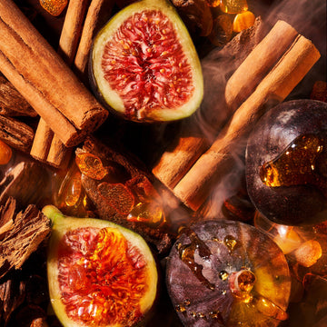 A close-up of halved and whole figs with cinnamon sticks and amber stones evokes the luxurious feel of Roja Parfums Amber Aoud Coffret. Warm lighting highlights rich textures, while a smoky haze creates a cozy atmosphere, akin to unwrapping an exquisite gift box.