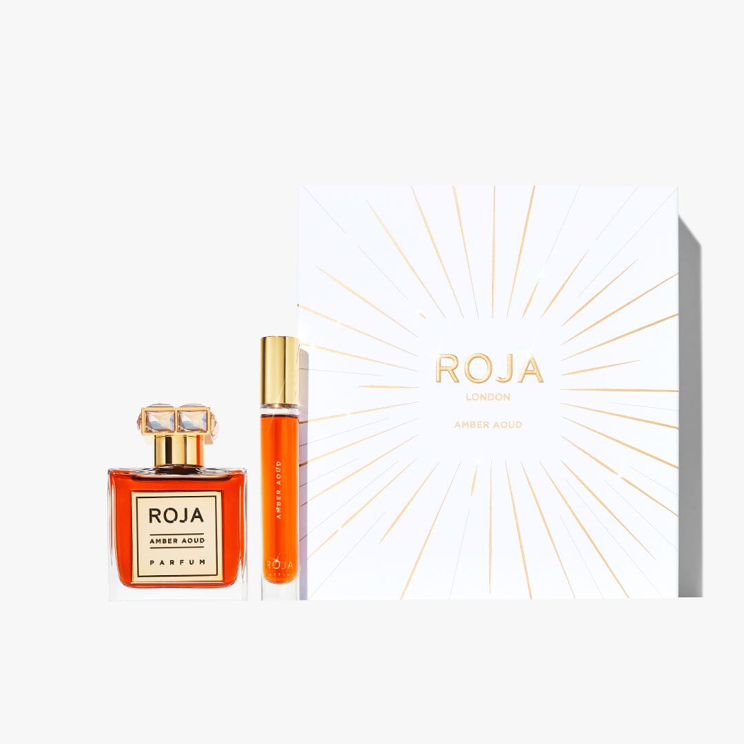 The Amber Aoud Festive Gift Set by Roja Parfums includes a square glass bottle and slim travel spray, both filled with orange liquid, beside a white box with gold rays—ideal for fans of rich Amber Aoud and oud notes.