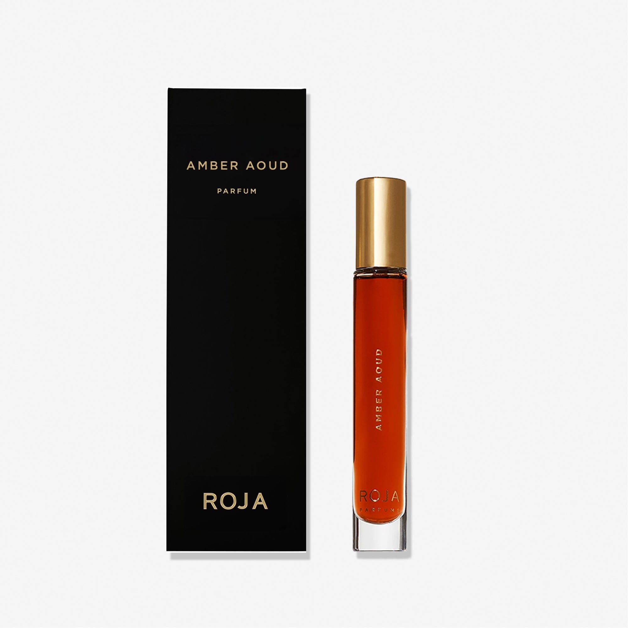 A tall, slim glass bottle of Roja Parfums Amber Aoud with a gold cap stands beside a sleek black box adorned with gold text—an elegant display against a plain white background.