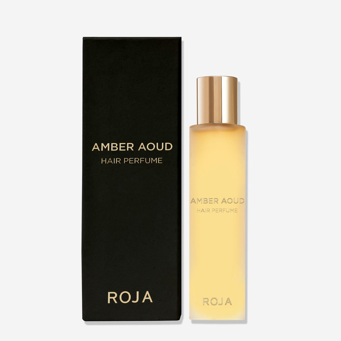 A Roja Parfums Amber Aoud Hair Perfume with a gold cap sits beside its black, gold-lettered box, both displayed on a light background. The fragrance elegantly blends rose and oud notes.