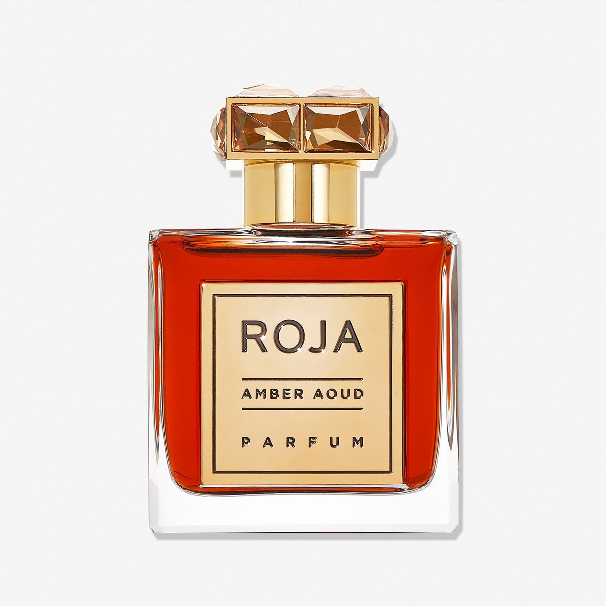 A clear glass bottle of Roja Parfums Enigma Aoud, an elegant aoud perfume with amber liquid, features a gold label and an ornate gold cap with gem-like details, set against a white background.