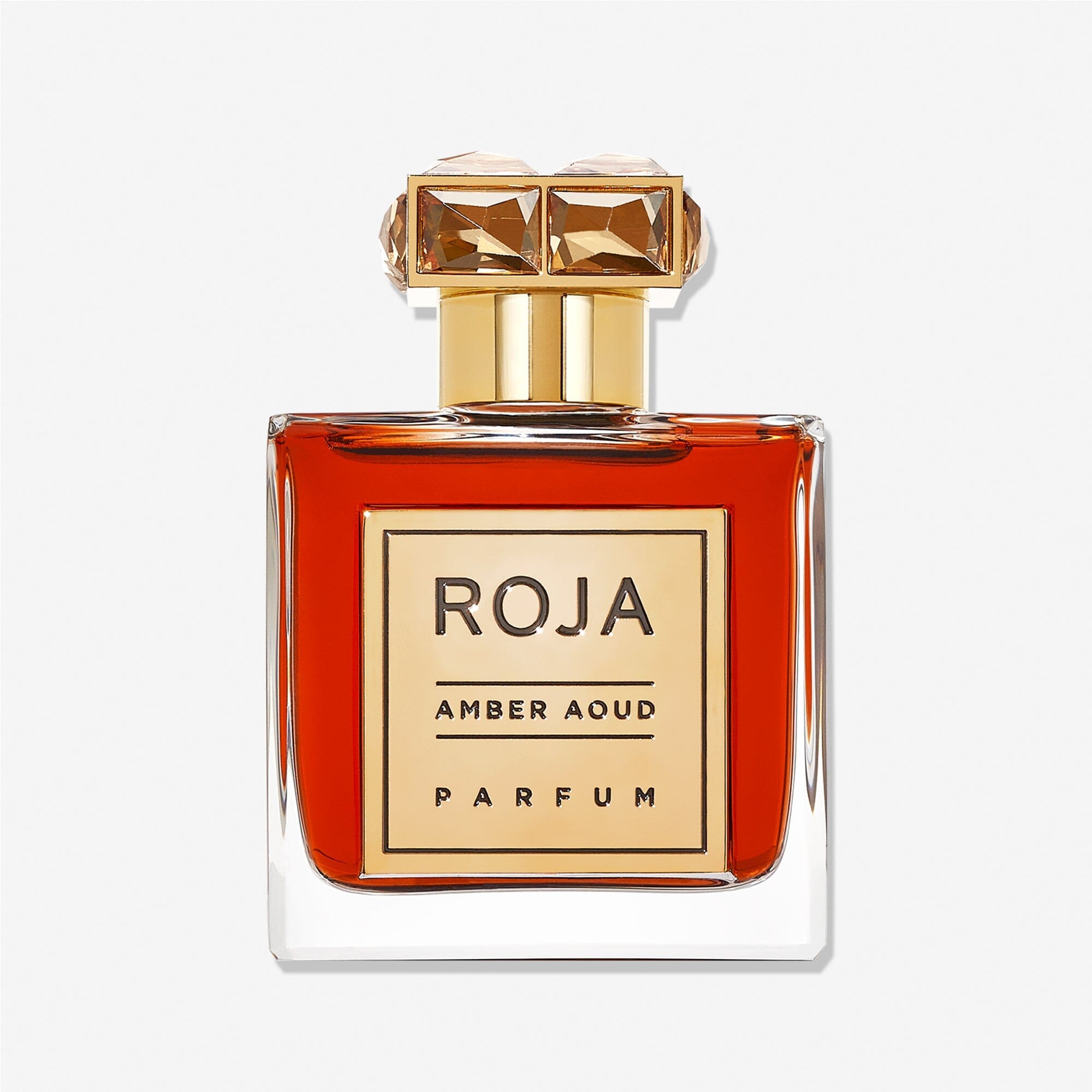 A clear glass bottle of Roja Parfums Enigma Aoud, an elegant aoud perfume with amber liquid, features a gold label and an ornate gold cap with gem-like details, set against a white background.