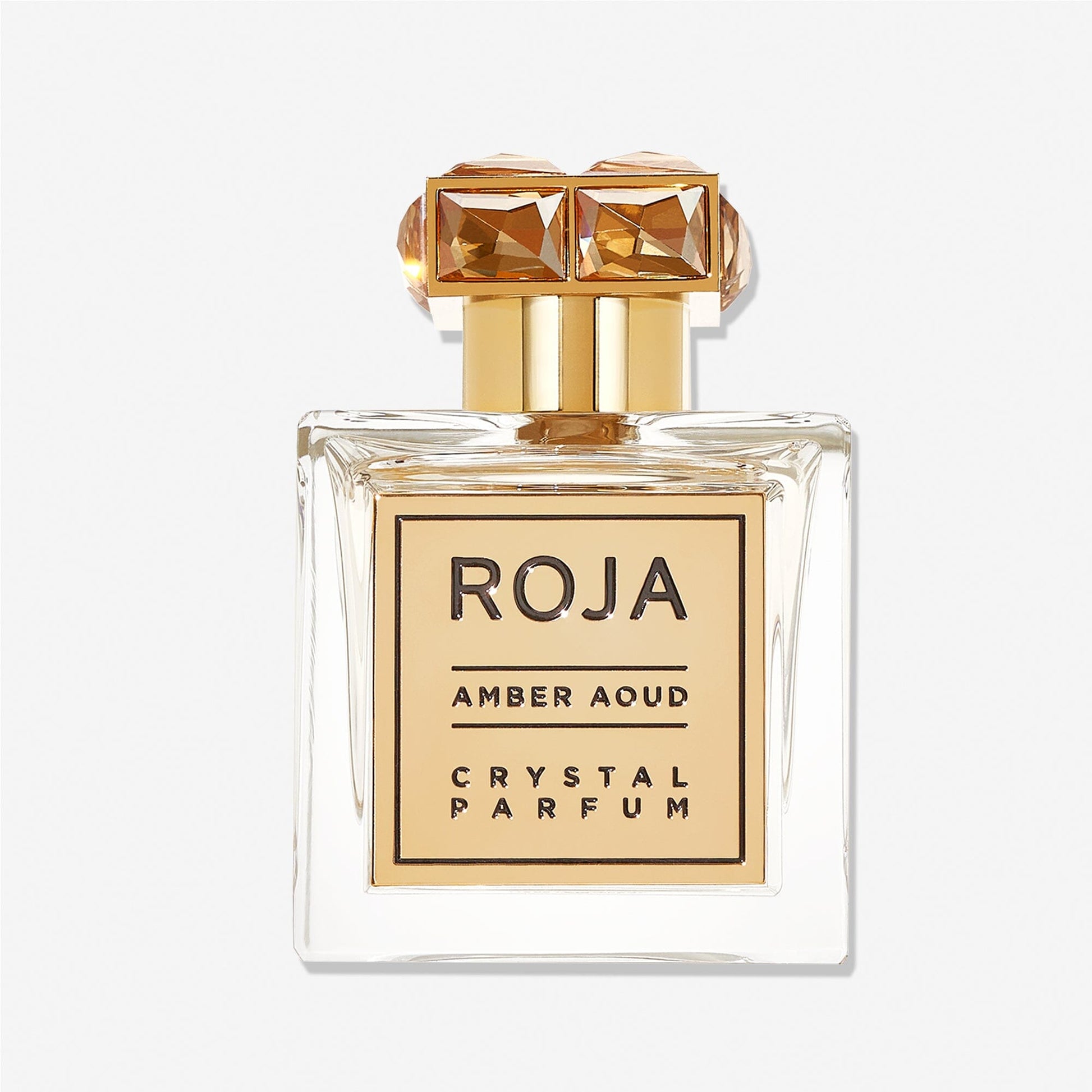 A clear glass bottle of Roja Parfums Aoud CRYSTAL™️ features a gold cap with two gem-like squares and a gold label with bold black text. The parfum blends rich Aoud and rose notes for an opulent fragrance experience.