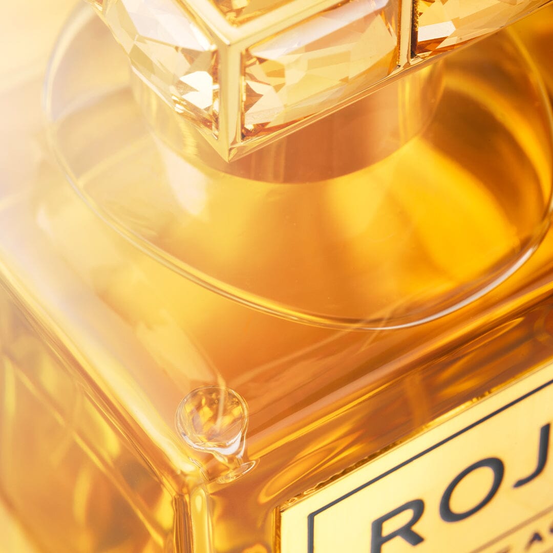 Close-up of an ornate perfume bottle with amber liquid, exuding luxury. The gem-like cap and elegant design reveal Aoud Extraordinaire by Roja Parfums. Warm lighting enhances its golden hues, capturing the essence of this exquisite oud fragrance.