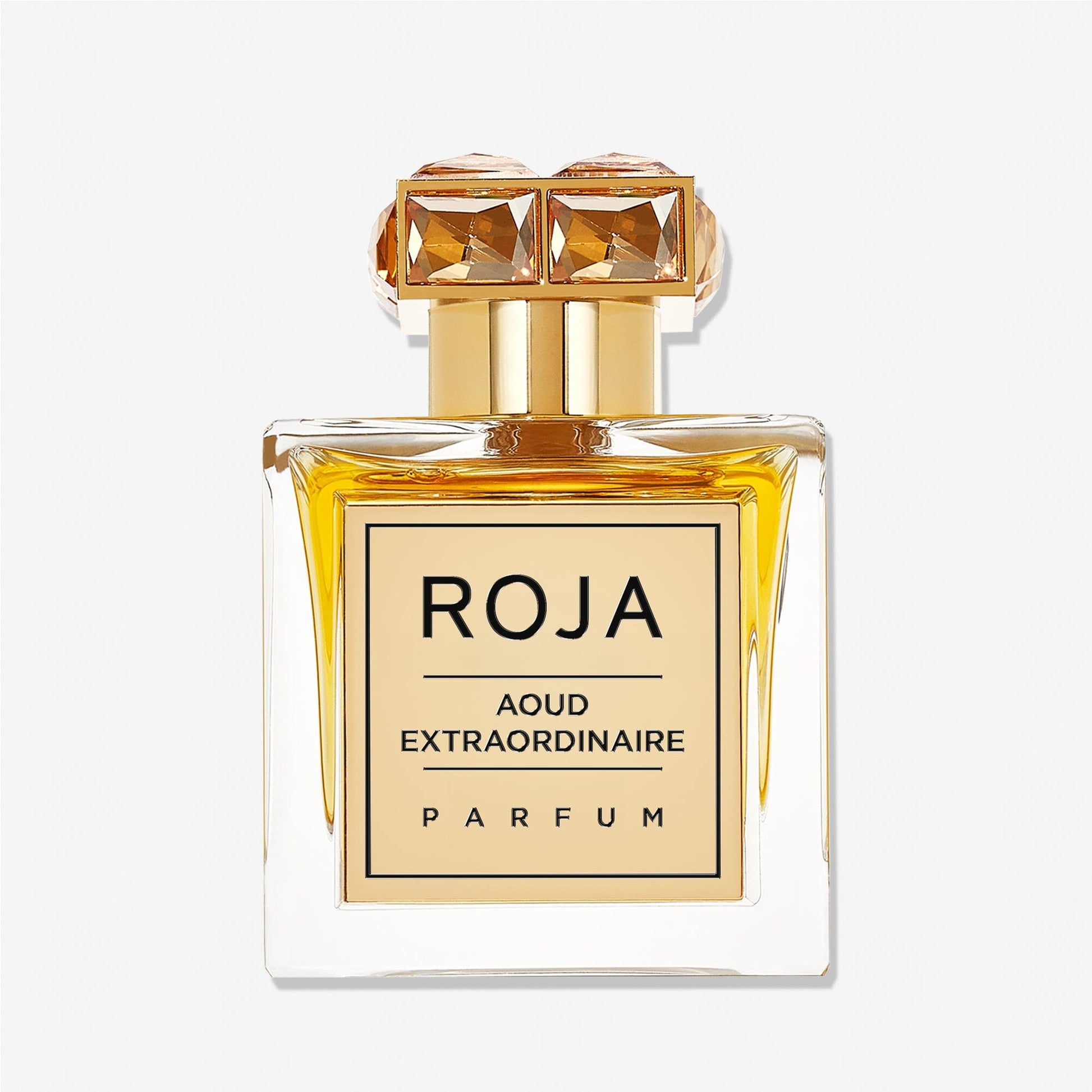 A square glass bottle of Roja Parfums Aoud Extraordinaire holds golden-yellow liquid, featuring a gold gem-accented cap and a front label reading AOUD EXTRAORDINAIRE, capturing the essence of refined oud.