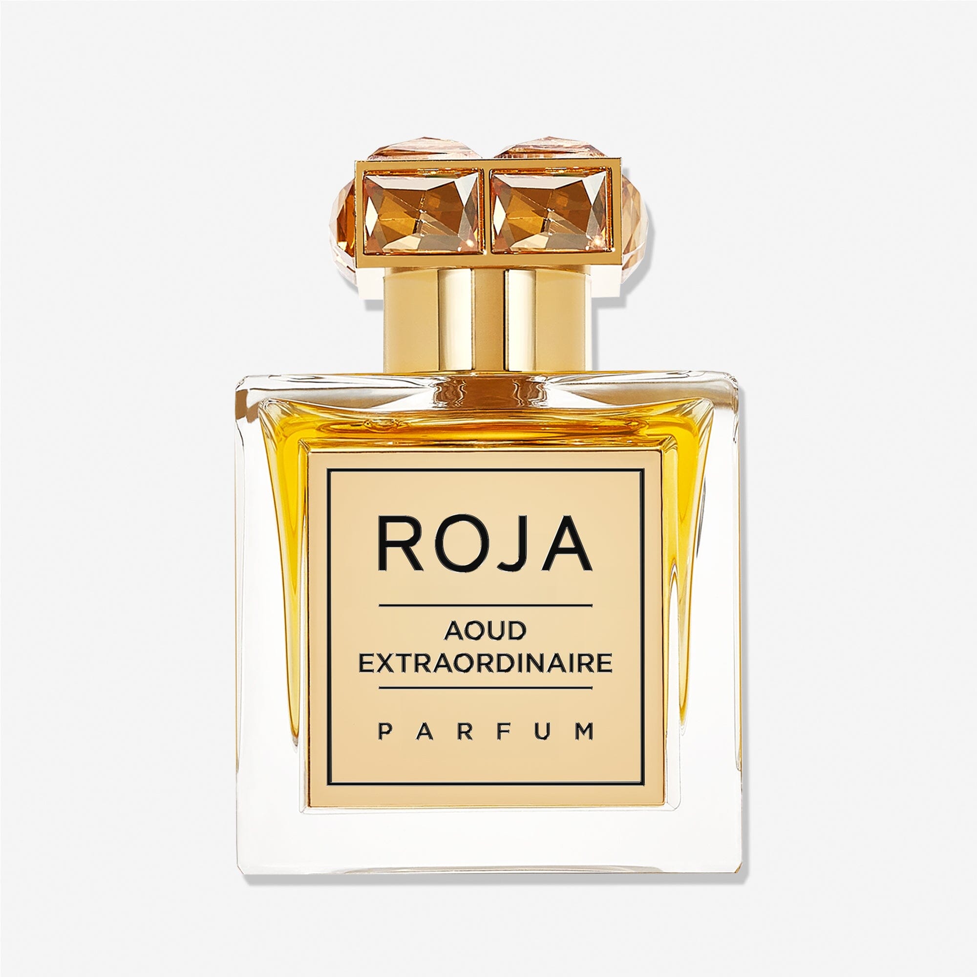 A square glass bottle of Roja Parfums Aoud Extraordinaire holds golden-yellow liquid, featuring a gold gem-accented cap and a front label reading AOUD EXTRAORDINAIRE, capturing the essence of refined oud.