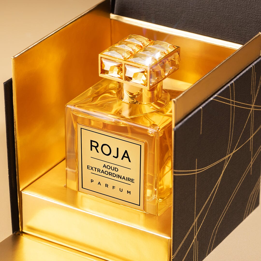 A lavish Roja Parfums Aoud Extraordinaire perfume bottle rests inside a black and gold box, exuding opulence. The transparent bottle with its gold cap beautifully reflects light, capturing the essence of Ouds remarkable allure.