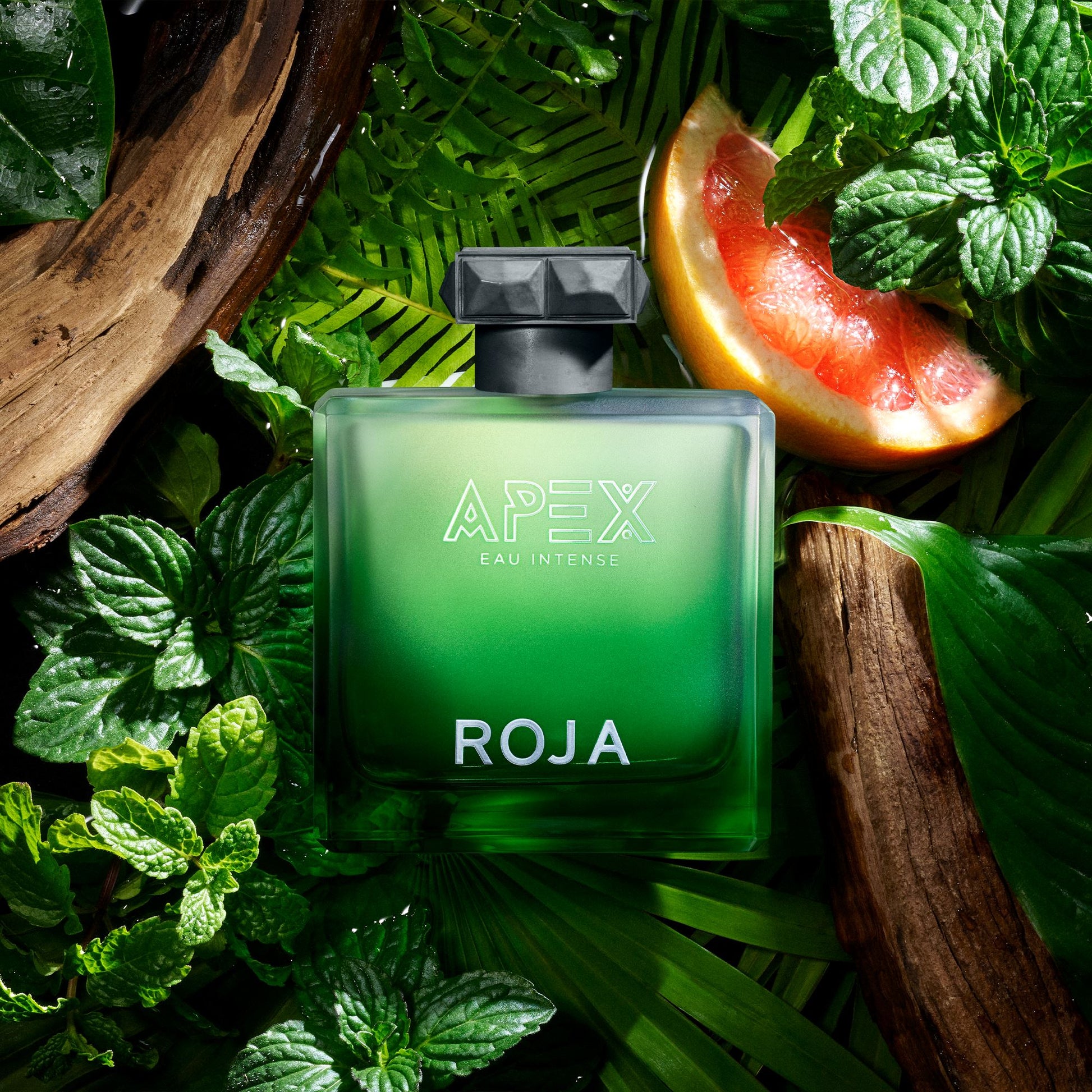 A green glass bottle of Roja Apex Eau Intense perfume sits among fresh mint leaves, a grapefruit slice, and pieces of wood, creating a lush, tropical atmosphere.