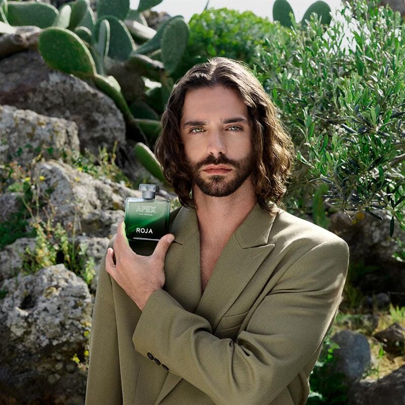 A bearded man with long brown hair in a tan suit holds a green bottle of Roja Parfums Apex Eau Intense outside among rocks, cacti, and greenery, capturing the bold essence of natural perfumery.
