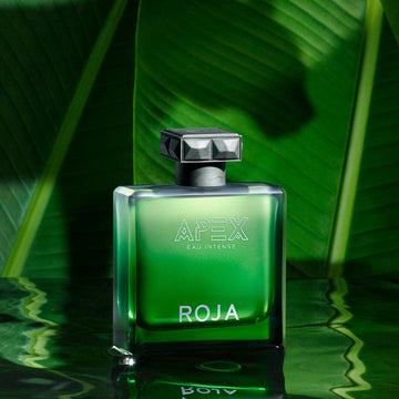 A green glass bottle of Apex Eau Intense by Roja Parfums rests on a reflective surface, inviting you to an olfactory adventure, with lush tropical foliage behind and a subtle Chypré accord lingering in the air.