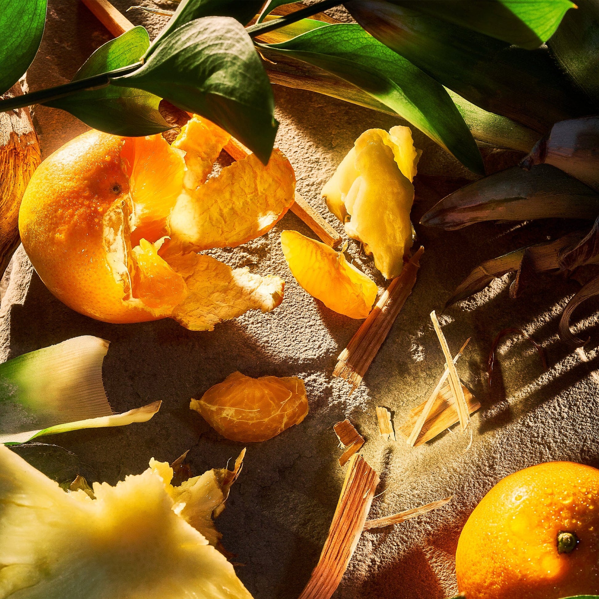 A close-up of peeled and whole oranges alongside pineapple pieces on a textured surface, partially covered by green leaves, casts dramatic shadows. This vibrant citrus blast adds freshness to the composition of Apex Pour Homme by Roja Parfums.