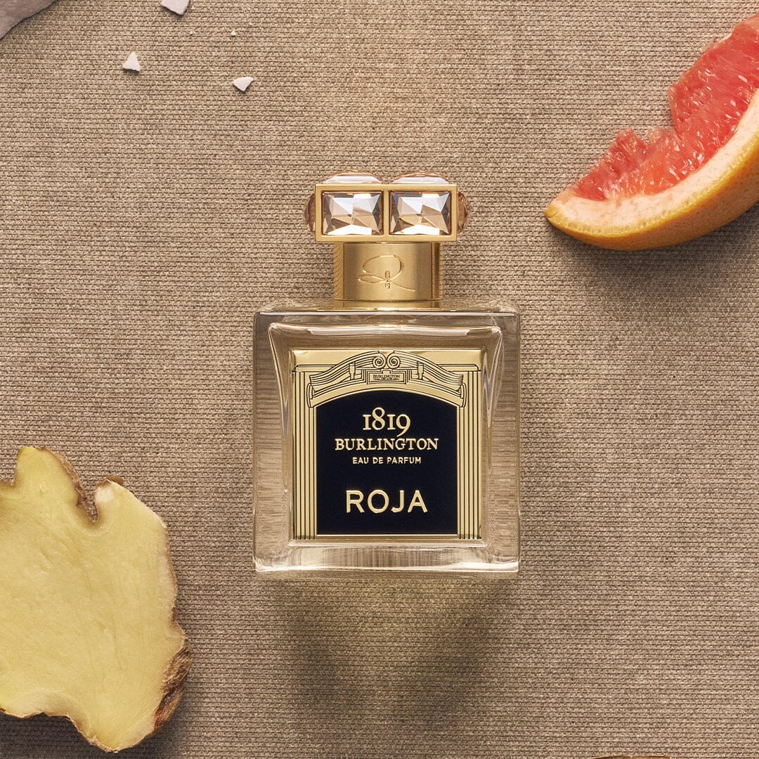 A bottle of Burlington 1819 by Roja Parfums rests on a textured brown surface, accompanied by a slice of ginger on the left and grapefruit on the top right—reflecting the essence of Burlington Arcade's premium fragrances.