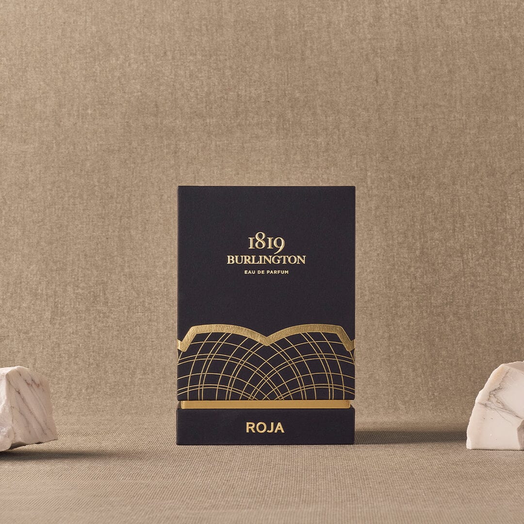 A dark blue and gold box labeled Burlington 1819 by Roja Parfums stands upright on a beige textured surface, flanked by two white marble pieces—reflecting the luxurious essence of Roja Parfums and the heritage of Burlington Arcade.