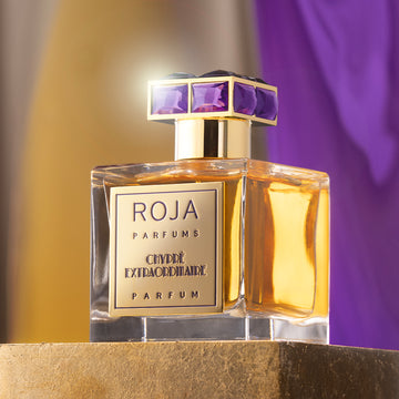 A bottle of Roja Parfums Cuvée Extraordinaire perfume with golden liquid, a gold label, and a gold cap adorned with large purple gems, set against a blurred yellow and purple background.