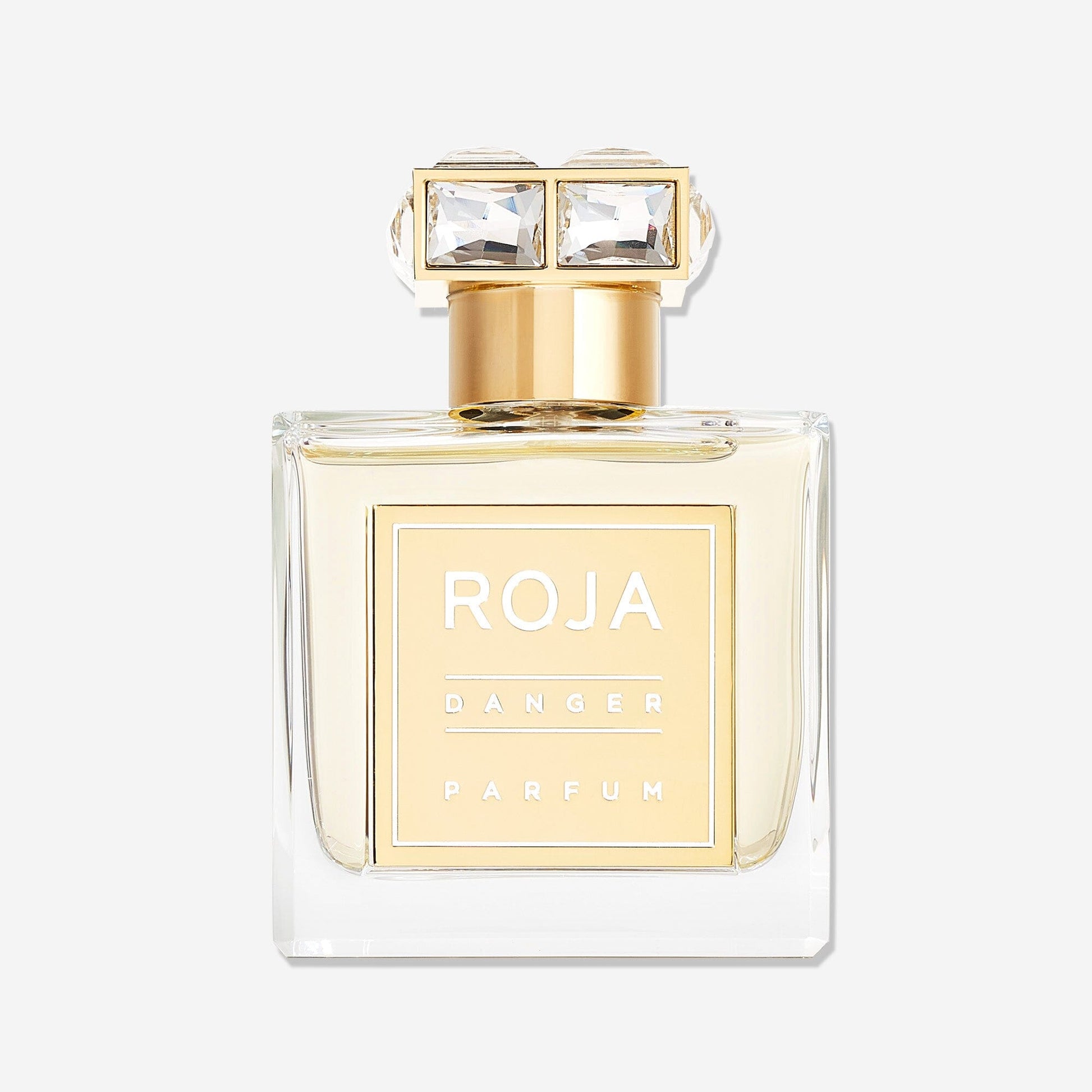 A clear rectangular perfume bottle with a gold label reading "Danger Pour Femme" by Roja Parfums and a gold cap adorned with two large square gemstones, the striking aphrodisiac fragrance is set against a plain white background.