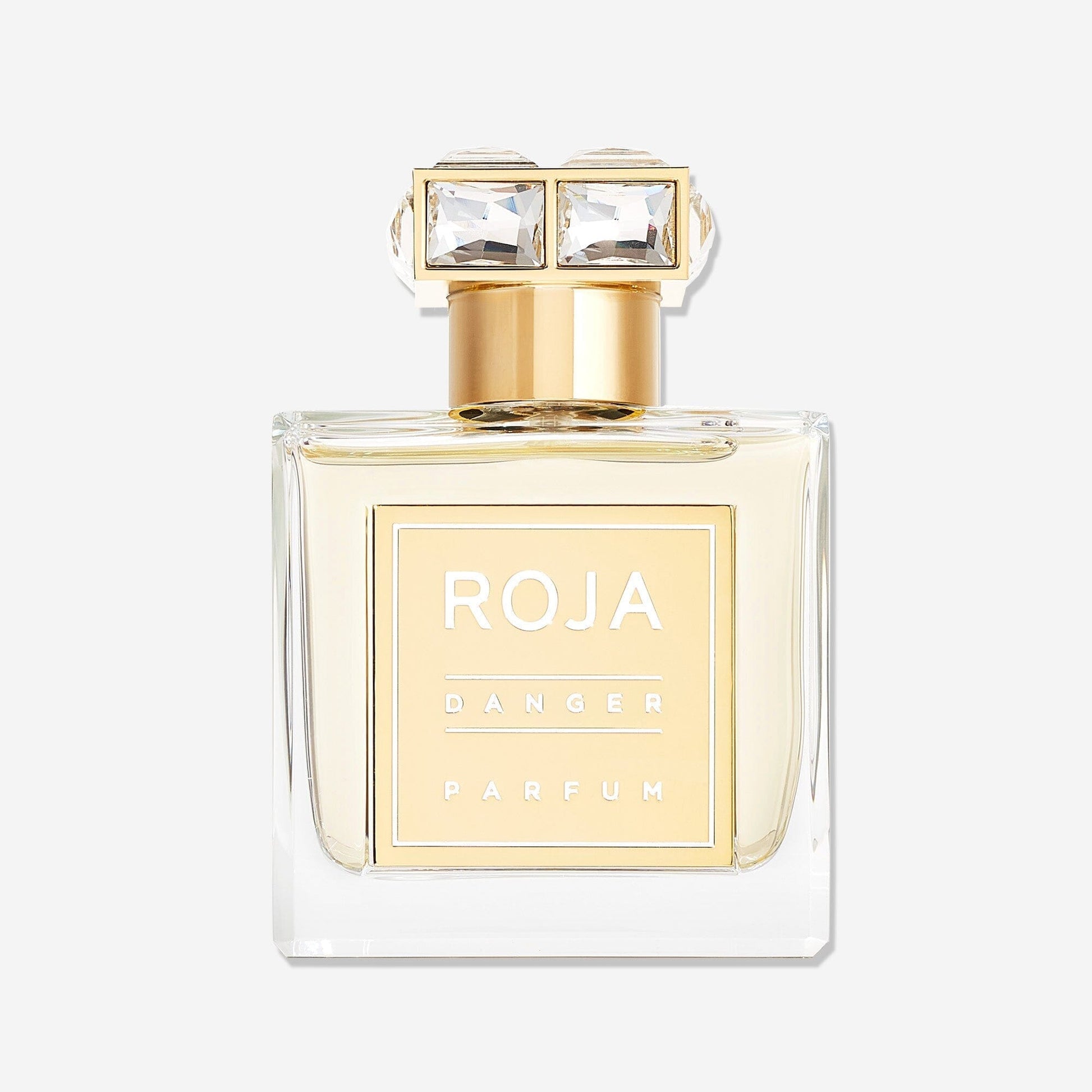 A clear glass bottle of Roja Parfums Danger Pour Femme, an aphrodisiac fragrance, features gold accents, a square gold label, and a gold cap with two large square gemstones. The bottle is shown against a plain white background.