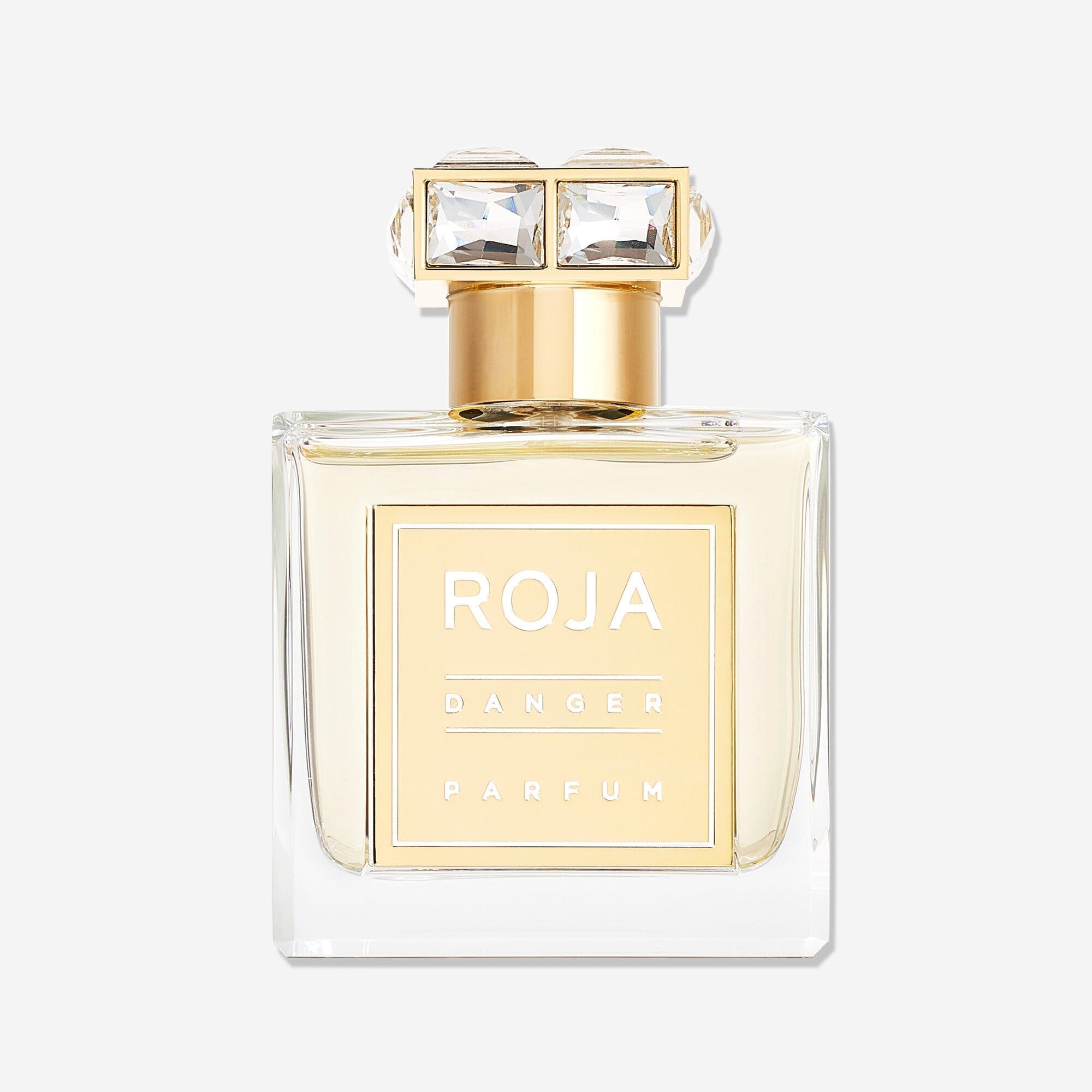 A clear glass bottle of Roja Parfums Danger Pour Femme, an aphrodisiac fragrance, features gold accents, a square gold label, and a gold cap with two large square gemstones. The bottle is shown against a plain white background.
