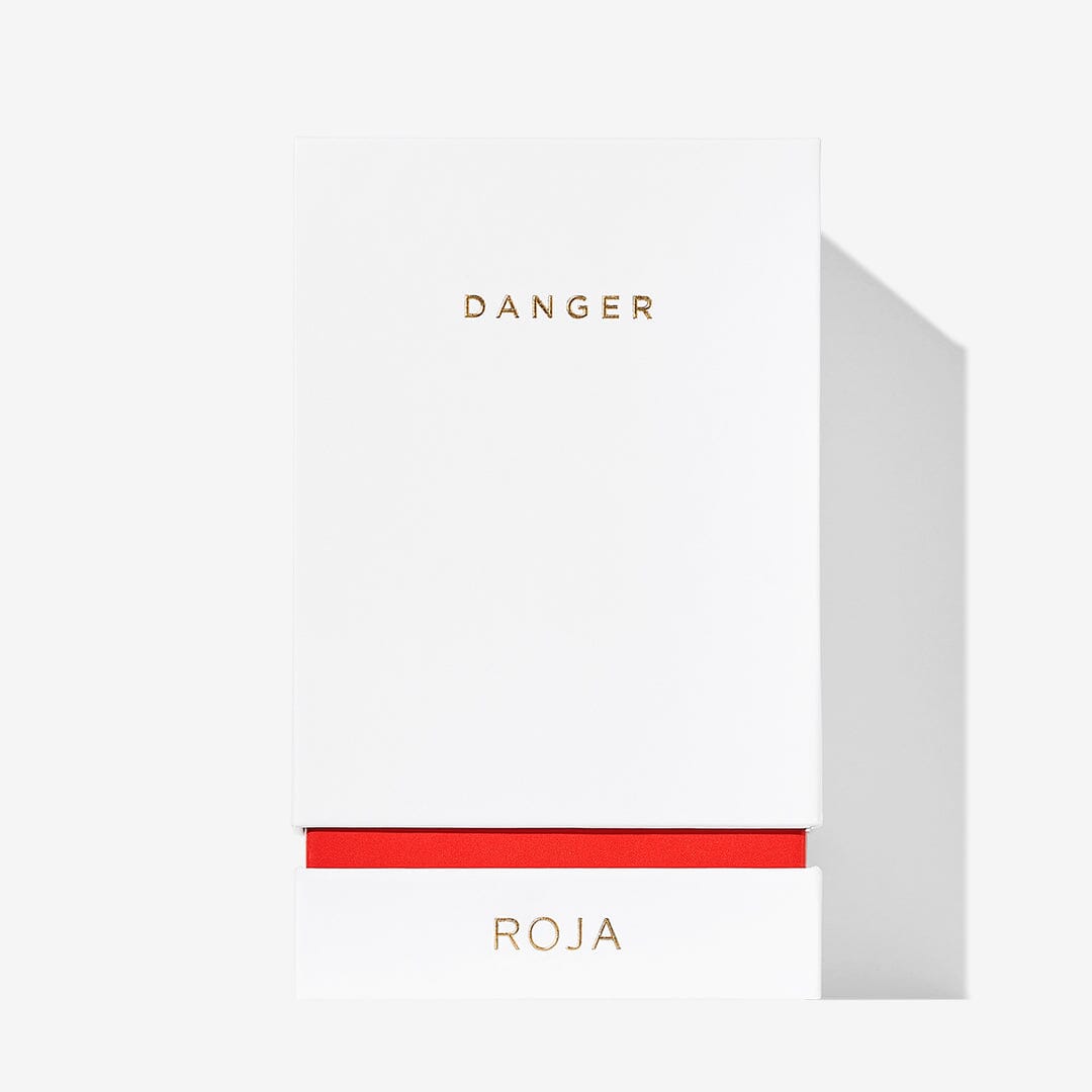 A minimalistic white box features gold text: DANGER at the top and ROJA PARFUMS at the bottom, divided by a red band. This hints at the alluring, aphrodisiac scent of Danger Pour Femme by Roja Parfums. Plain white background.