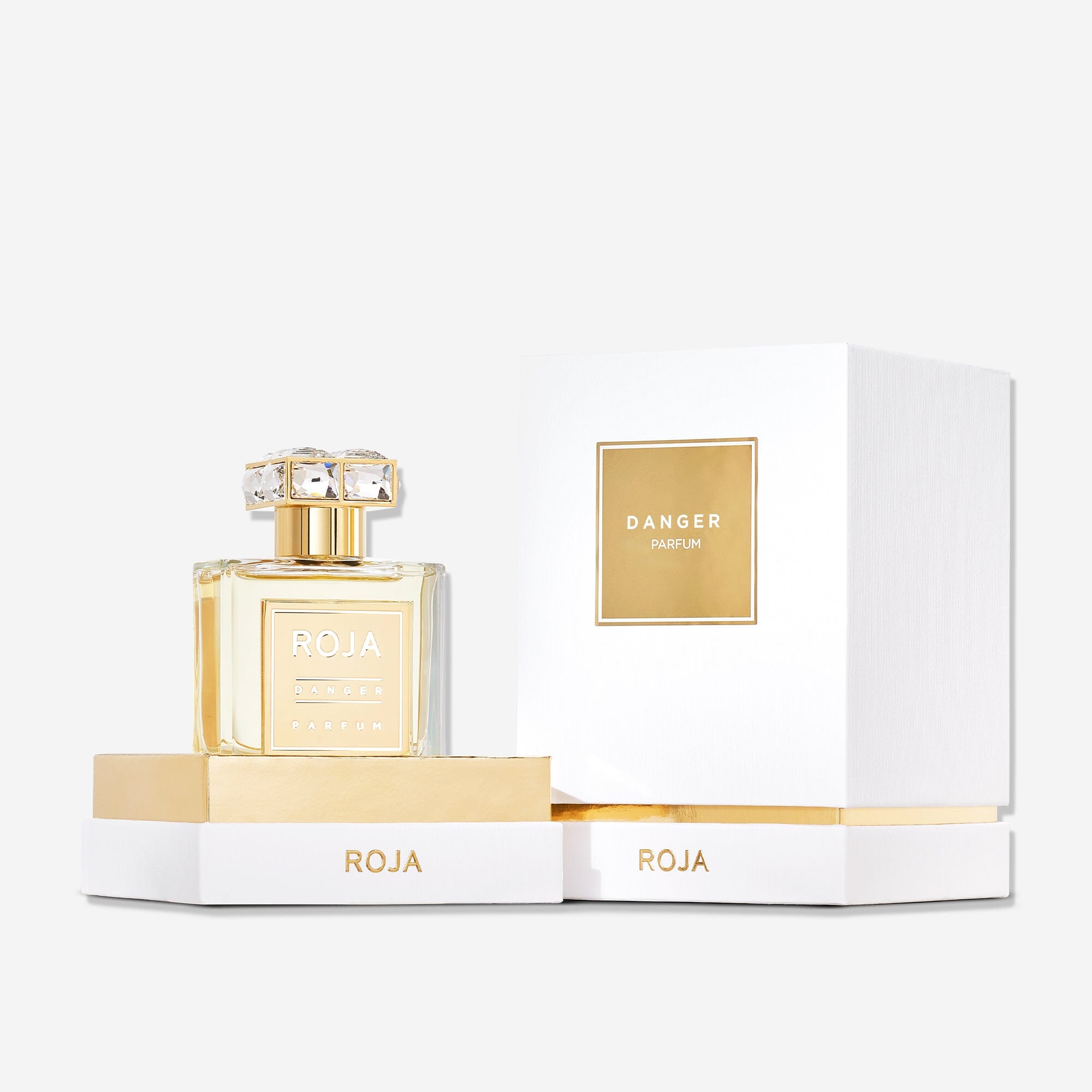 A bottle of Danger Pour Femme by Roja Parfums sits before its elegant white and gold box, the jeweled cap placed behind. The clear bottle displays a bold gold label, hinting at its femme fatale scent.