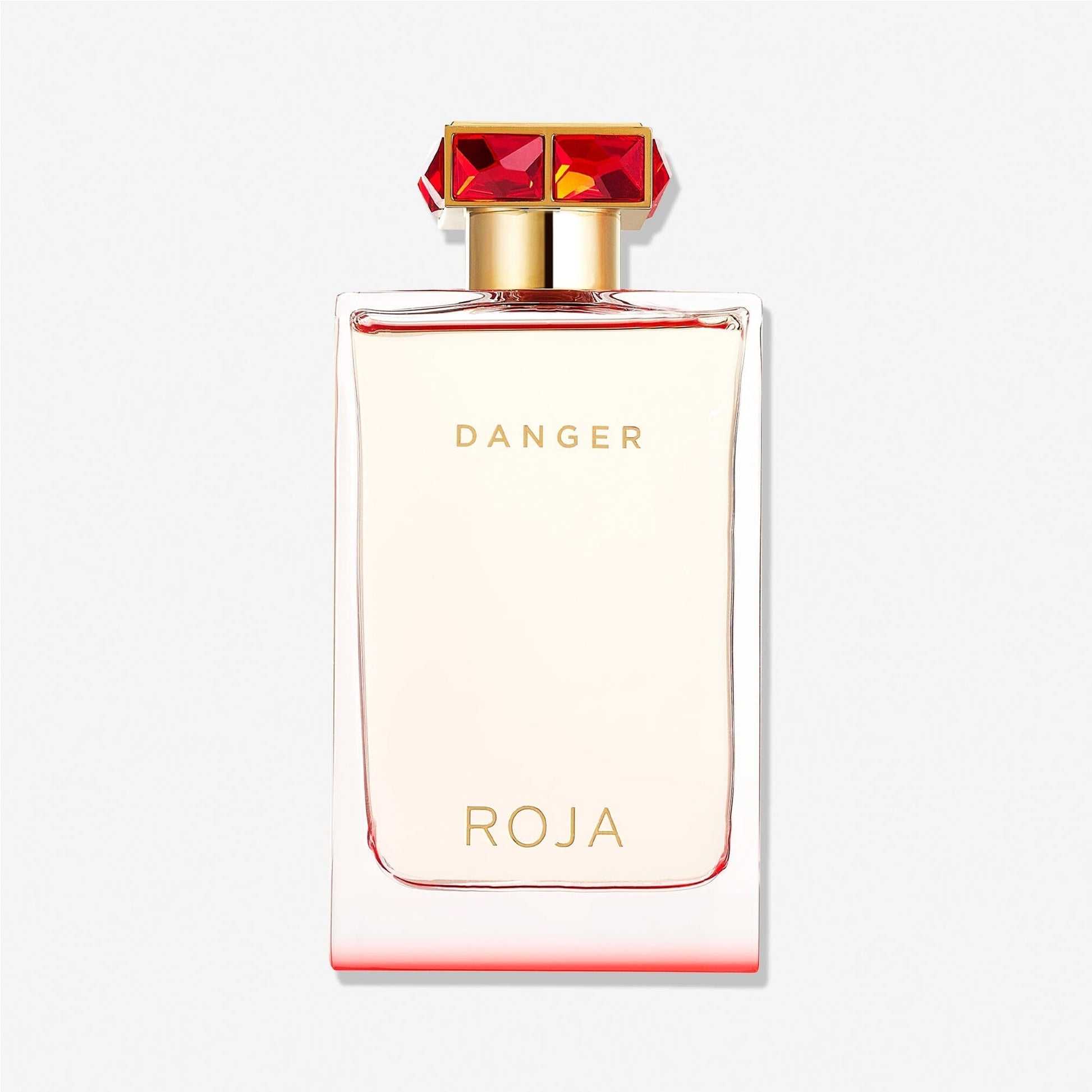 A clear rectangular bottle labeled "Danger Pour Femme" and "Roja Parfums" in gold, topped with a gold cap adorned with red gemstones, suggests an aphrodisiac fragrance against a plain white background.