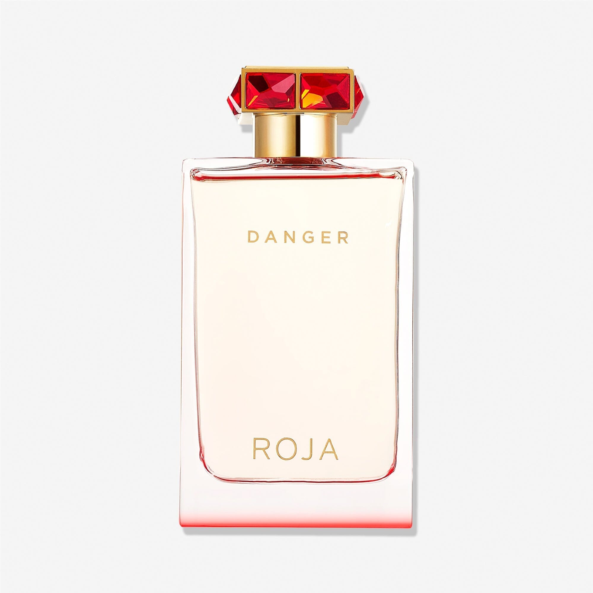 A clear rectangular bottle labeled "Danger Pour Femme" and "Roja Parfums" in gold, topped with a gold cap adorned with red gemstones, suggests an aphrodisiac fragrance against a plain white background.