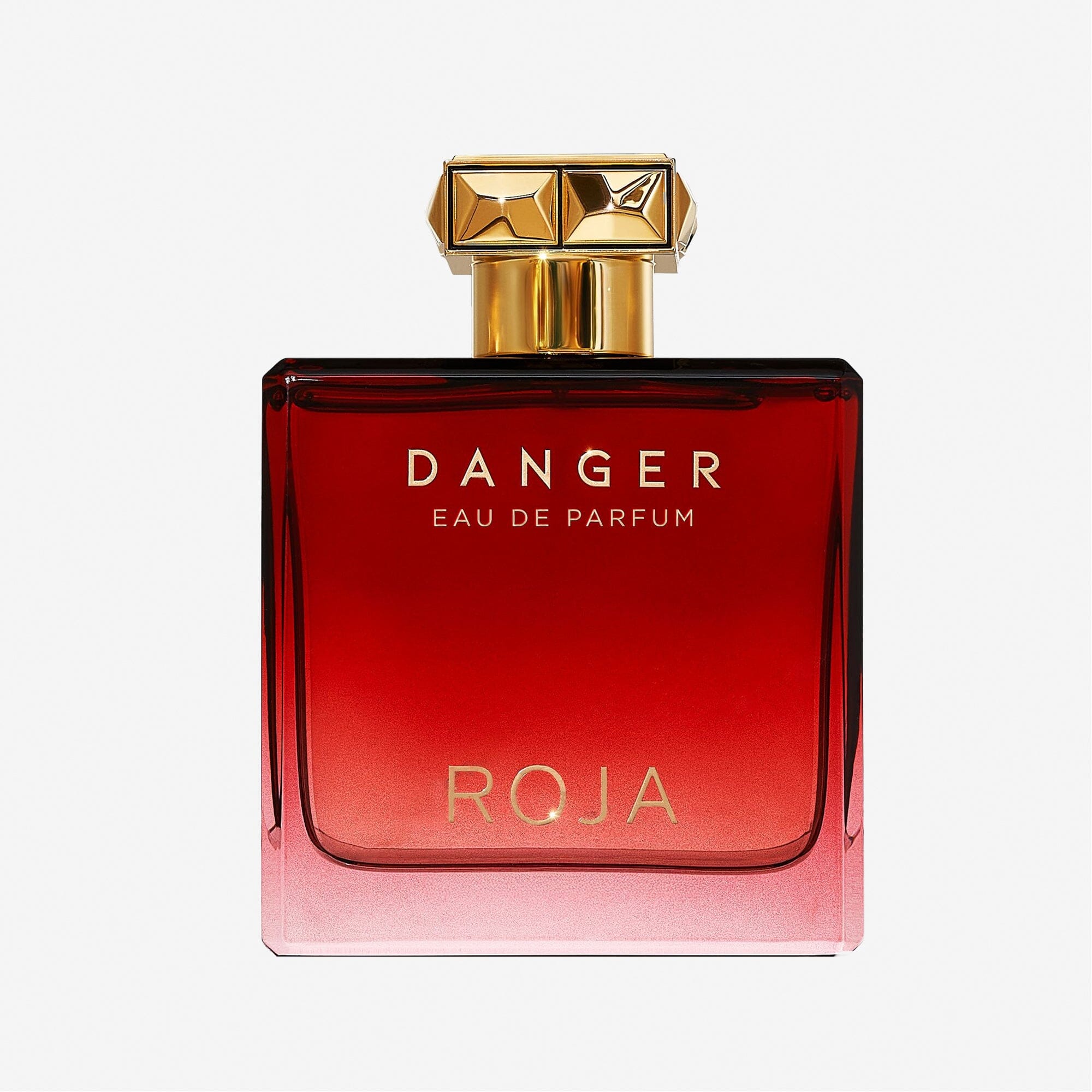A square glass bottle of Roja Parfums Danger Pour Homme, featuring a red and gold label and a gold geometric cap, showcases the seductive mens fragrance against a plain white background.