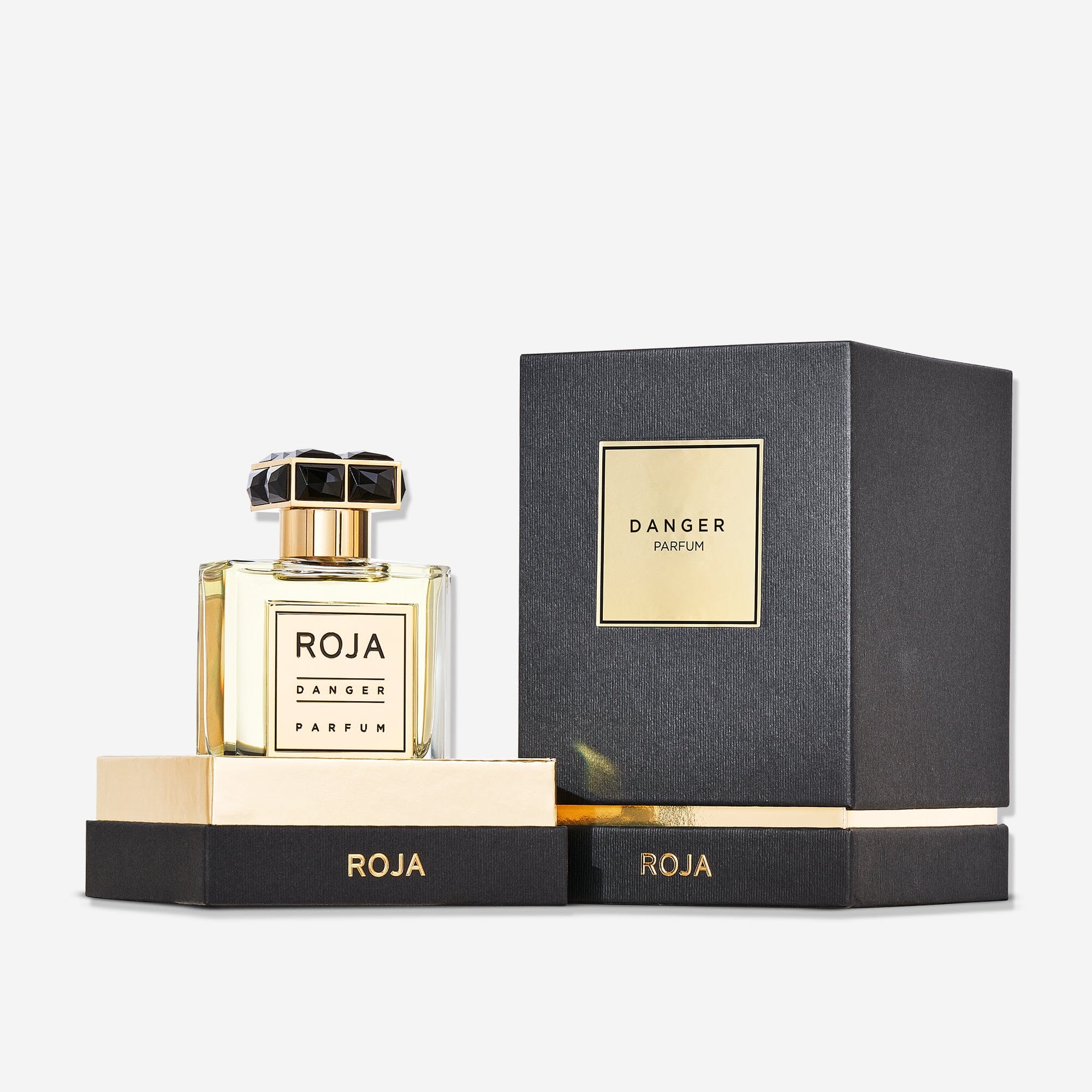 A bottle of Danger Pour Homme by Roja Parfums, with notes of lavender and patchouli, rests beside its black and gold box on matching black bases with gold accents, featuring the brand name ROJA in gold letters.