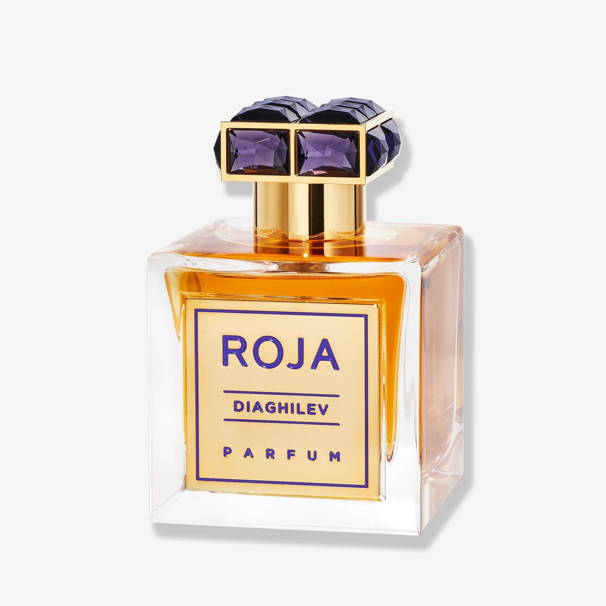 A clear glass bottle of Diaghilev by Roja Parfums holds amber liquid, gold accents, and a square gold label with purple text. The cap displays purple and gold geometric designs—an olfactory symphony of Chypré fragrance.