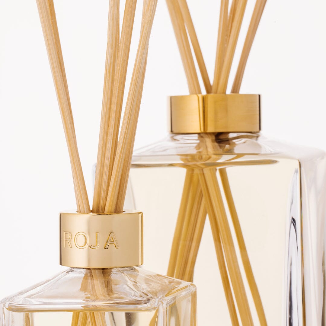 Two glass reed diffusers with gold caps and light wooden sticks, one engraved with ROJA London. The bottles contain the elegant "Lost in Paris" diffuser, filling your space with a light, gourmand fragrance.