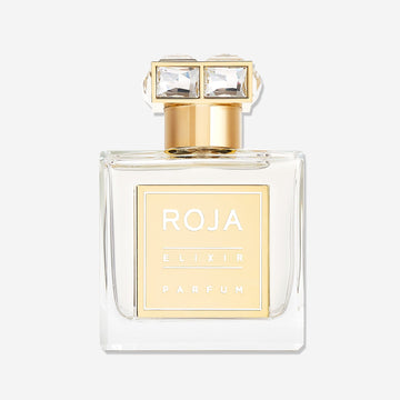A clear glass bottle of Roja Parfums Elixir Pour Femme with a gold label and cap, topped with two square-cut gemstones, set against a white background—an ethereal fragrance blending vanilla amber musk notes.